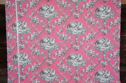 Pink fabric with a classic gray toile de Jouy pattern featuring peacocks, floral motifs, and a white measuring tape running vertically along the left edge, set against a dark wood background.