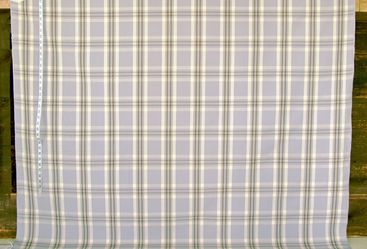 A close-up of a plaid fabric with a light gray base and intersecting cream, olive green, and tan stripes, shown flat with a measuring tape visible along the left edge.