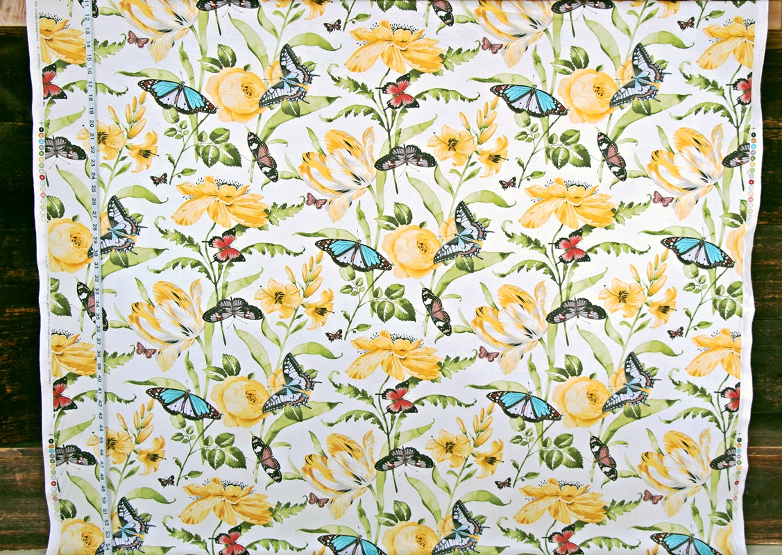 A white fabric panel or curtain featuring a detailed botanical and insect pattern with yellow flowers, green leaves, and blue butterflies, laid flat against a dark wood background.