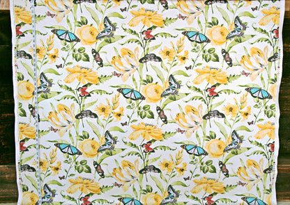 A white fabric panel or curtain featuring a detailed botanical and insect pattern with yellow flowers, green leaves, and blue butterflies, laid flat against a dark wood background.