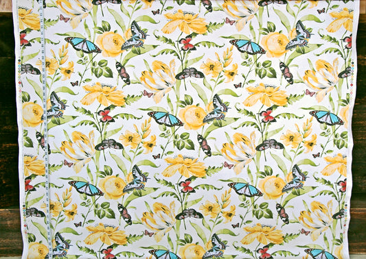 A white fabric panel or curtain featuring a detailed botanical and insect pattern with yellow flowers, green leaves, and blue butterflies, laid flat against a dark wood background.