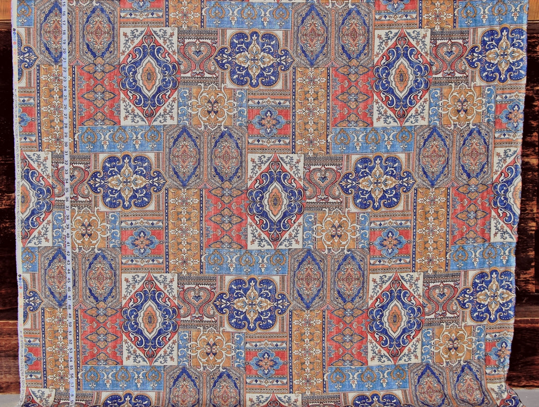 A close-up of a patterned textile or rug fabric with an intricate geometric and floral design in blue, rust red, beige, and brown tones, laid flat against a dark wood background.
