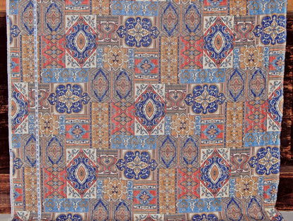 A close-up of a patterned textile or rug fabric with an intricate geometric and floral design in blue, rust red, beige, and brown tones, laid flat against a dark wood background.