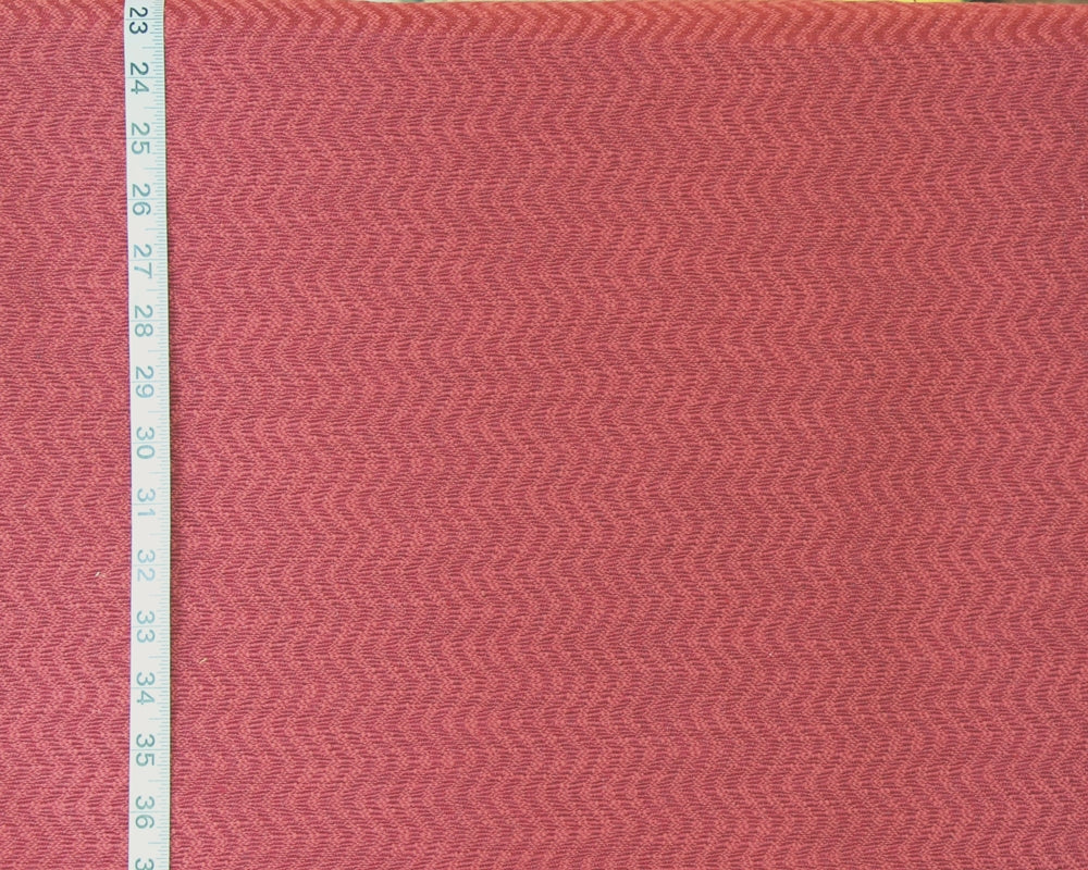 A close-up of a textured fabric with a pink and red herringbone pattern, shown next to a white measuring tape on the left side.