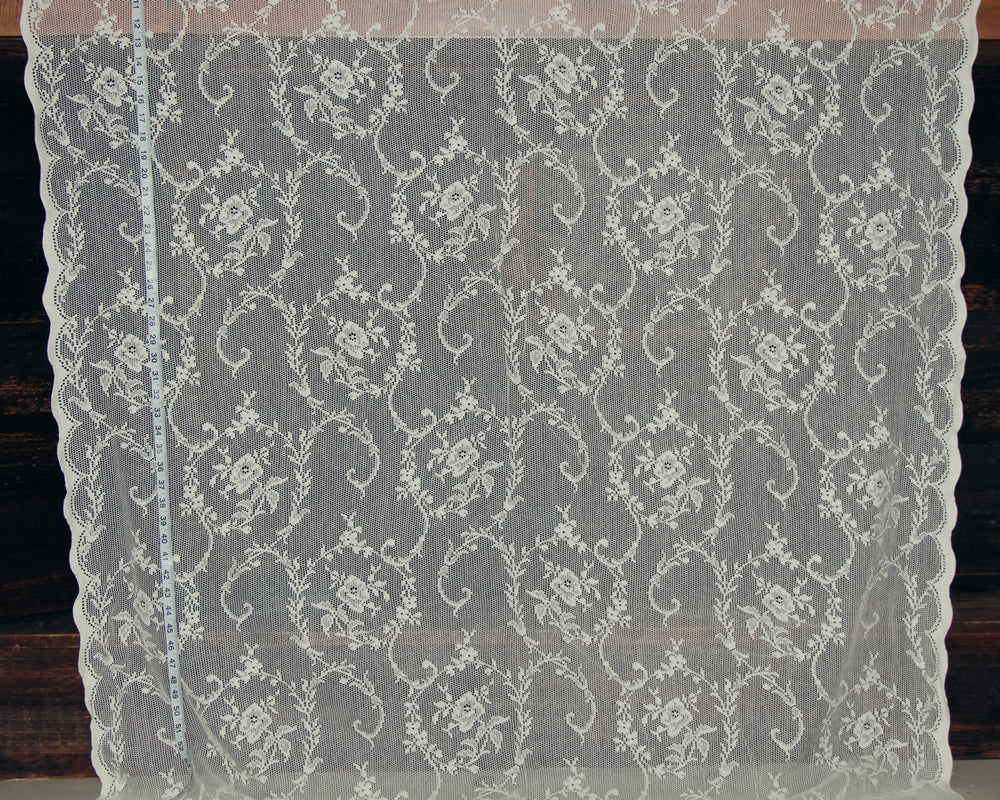 White floral lace fabric with a scalloped edge, displayed flat against a dark wood background with a measuring tape visible on the left side.