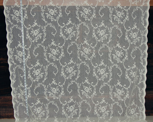 White floral lace fabric with a scalloped edge, displayed flat against a dark wood background with a measuring tape visible on the left side.