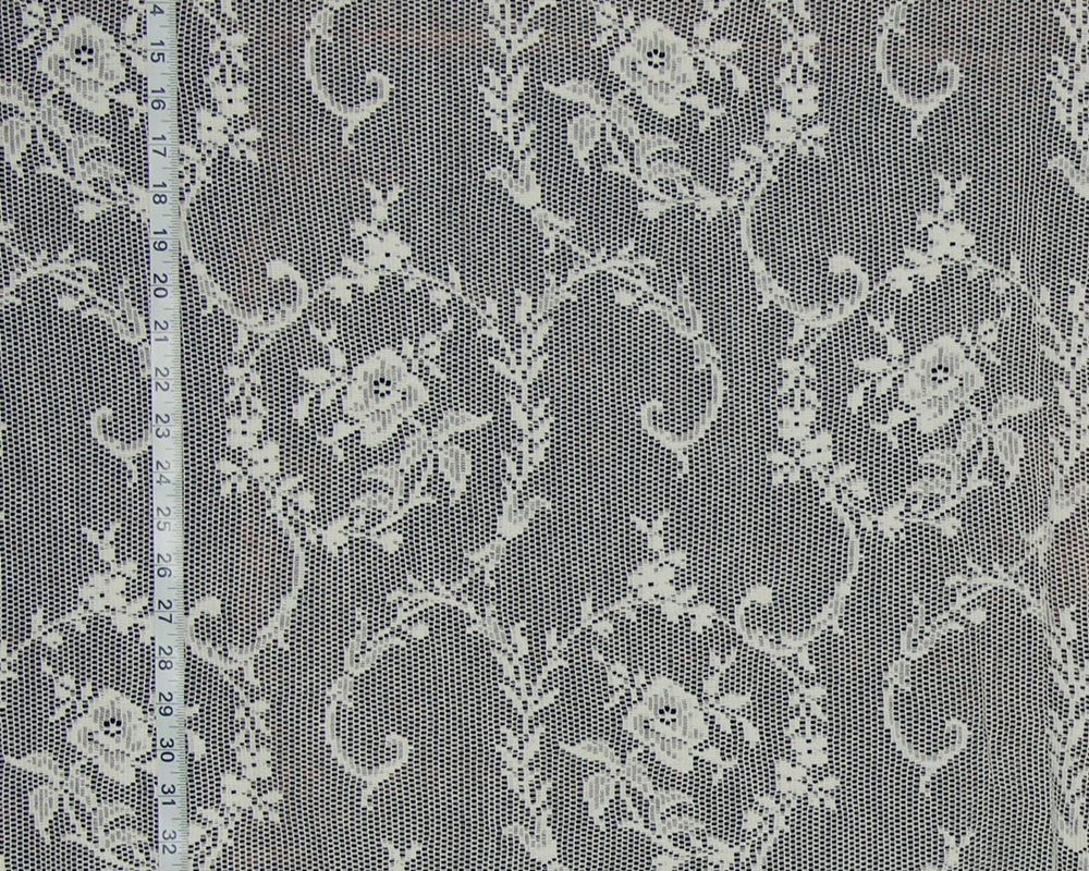 White floral lace fabric with a repeating damask pattern, shown flat with a white measuring tape on the left side for scale.