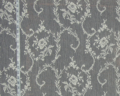 White floral lace fabric with a repeating damask pattern, shown flat with a white measuring tape on the left side for scale.