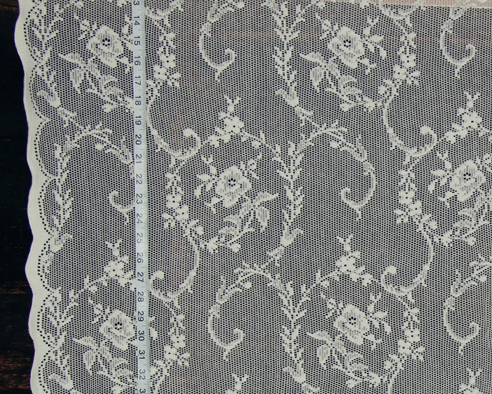 White floral lace fabric with a scalloped edge, shown flat against a dark background with a measuring tape running vertically to indicate scale.