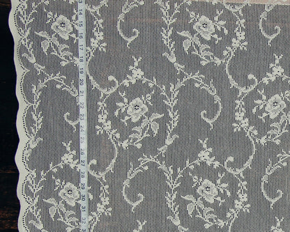 White floral lace fabric with a scalloped edge, shown flat against a dark background with a measuring tape running vertically to indicate scale.