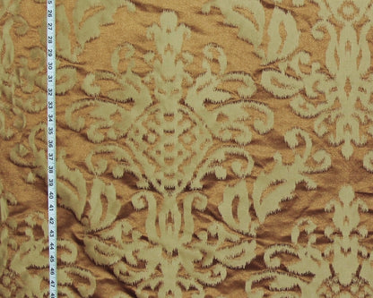 A close-up of a damask patterned fabric in warm tan and cream, with a white measuring tape visible along the left edge for scale.