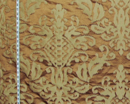 A close-up of a damask patterned fabric in warm tan and cream, with a white measuring tape visible along the left edge for scale.