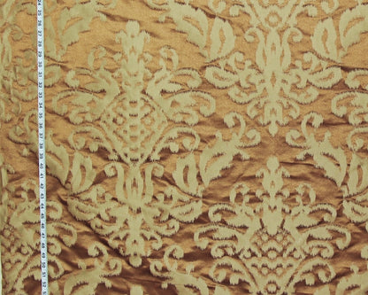 A close-up of damask patterned fabric in warm brown and beige, with a measuring tape visible along the left edge for scale.