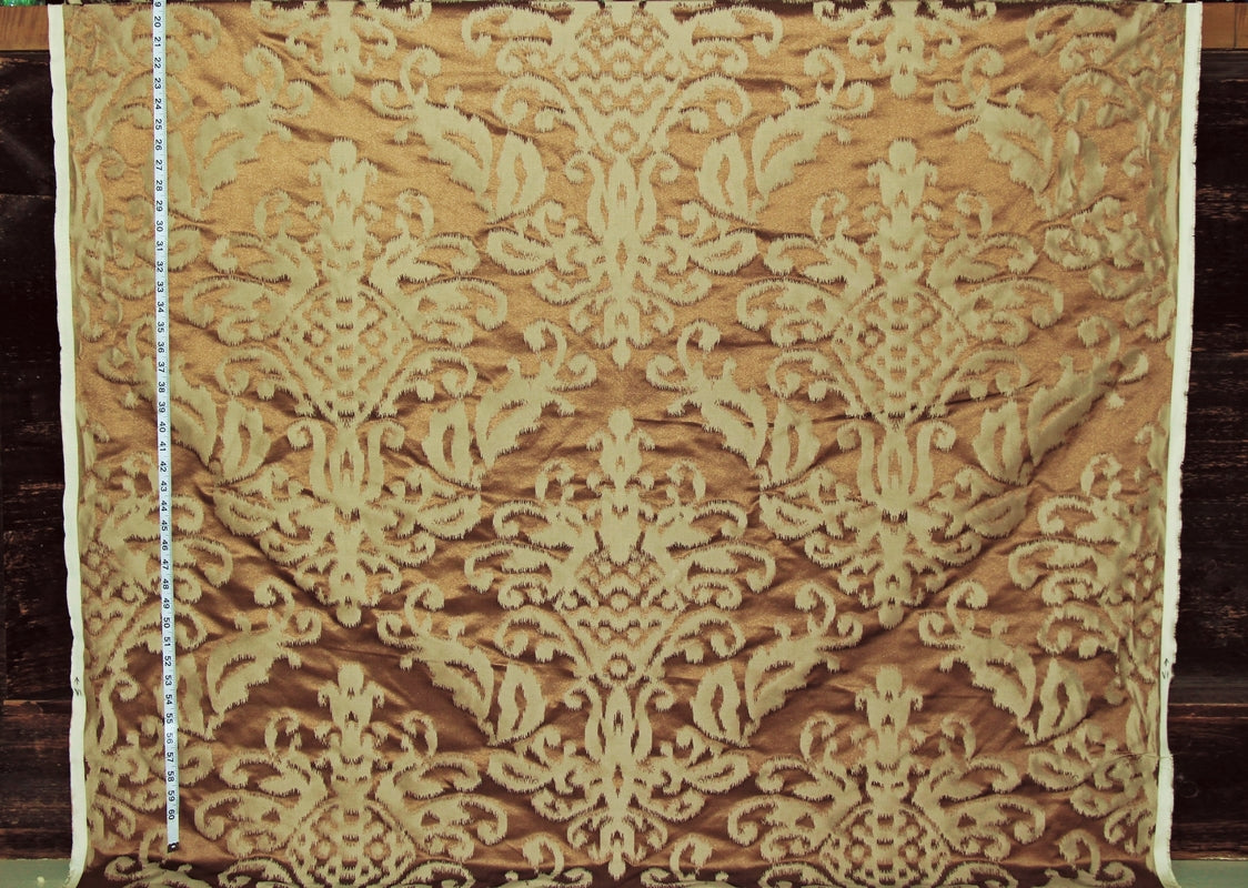 Damask patterned fabric in warm tan and beige, shown flat with a measuring tape along the left edge for scale.