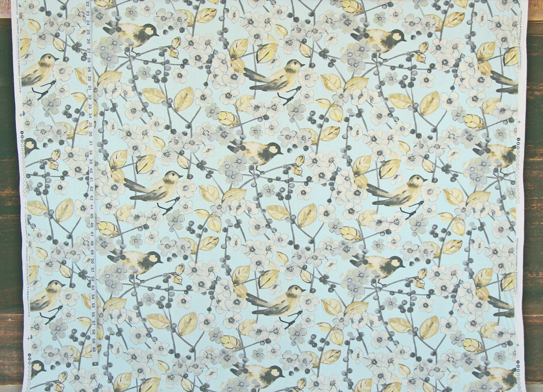 Fabric featuring a repeating pattern of small birds perched on branches with pale yellow leaves and berries on a light aqua background, laid flat on a dark wood surface.