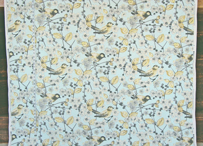 Fabric featuring a repeating pattern of small birds perched on branches with pale yellow leaves and berries on a light aqua background, laid flat on a dark wood surface.