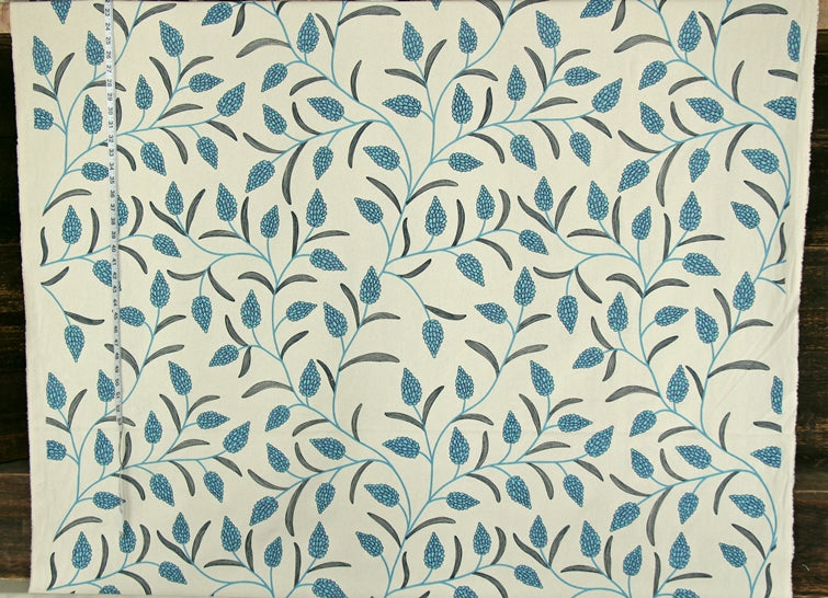A close-up of a fabric or wallpaper pattern featuring a repeating design of blue and gray leaves and vines on an off-white background, with a measuring tape visible along the left edge.