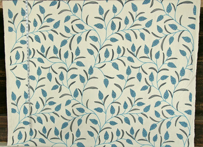 A close-up of a fabric or wallpaper pattern featuring a repeating design of blue and gray leaves and vines on an off-white background, with a measuring tape visible along the left edge.