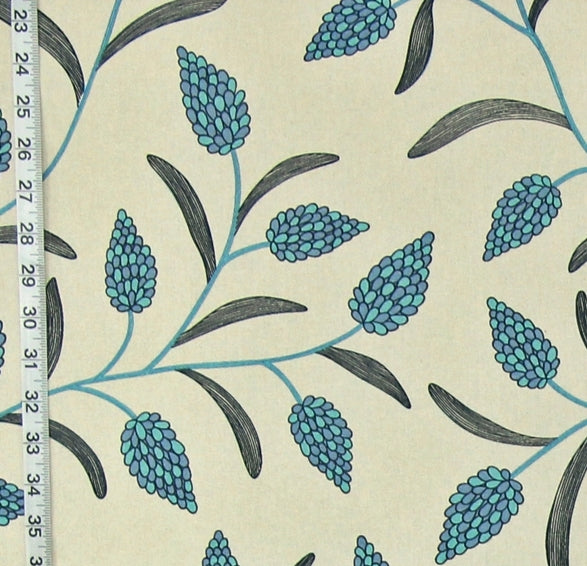 A close-up of a fabric or wallpaper pattern featuring blue-green floral buds and dark gray leaves on a warm cream background, with a white measuring tape visible along the left edge for scale.