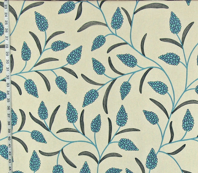 A close-up of a fabric pattern featuring teal-blue pinecones and dark gray leaves on a warm cream background, with a measuring tape visible along the left edge.