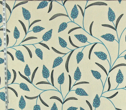 A close-up of a fabric pattern featuring teal-blue pinecones and dark gray leaves on a warm cream background, with a measuring tape visible along the left edge.