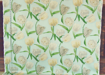 A close-up of a fabric or wallpaper pattern featuring a repeating design of tulips in cream, beige, and light gray on a pale mint green background, with a measuring tape visible along the left edge.