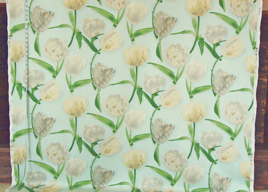 A close-up of a fabric or wallpaper pattern featuring a repeating design of tulips in cream, beige, and light gray on a pale mint green background, with a measuring tape visible along the left edge.