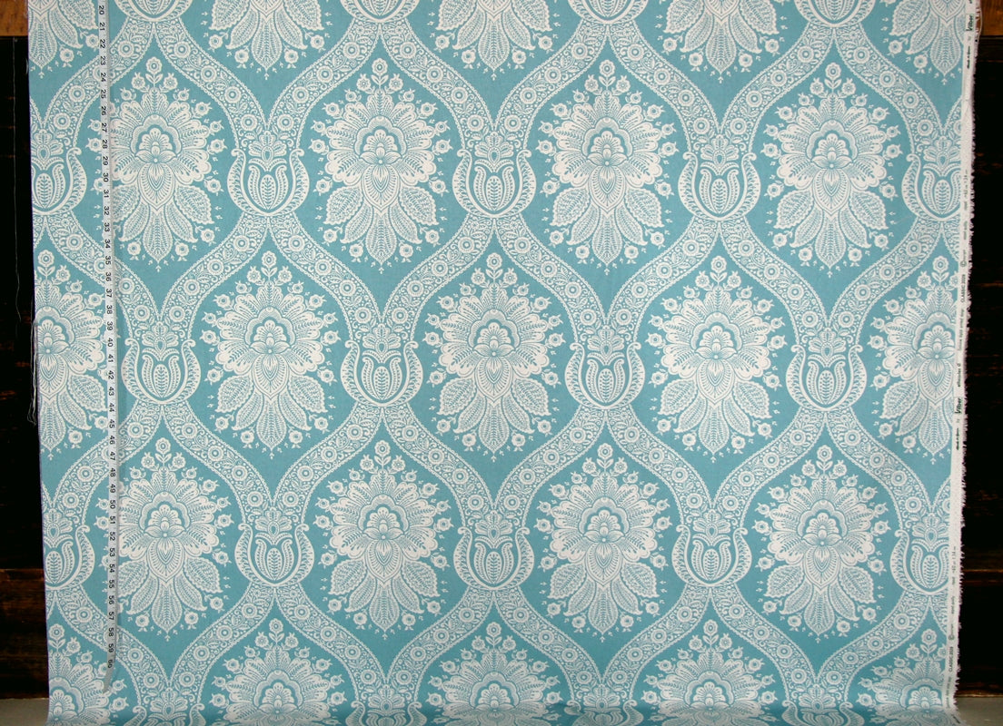 A close-up of a fabric or wallpaper sample with a repeating damask-style floral pattern in light aqua blue and white, shown against a dark background.