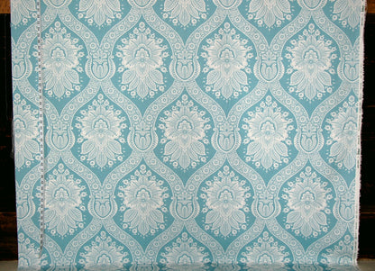 A close-up of a fabric or wallpaper sample with a repeating damask-style floral pattern in light aqua blue and white, shown against a dark background.