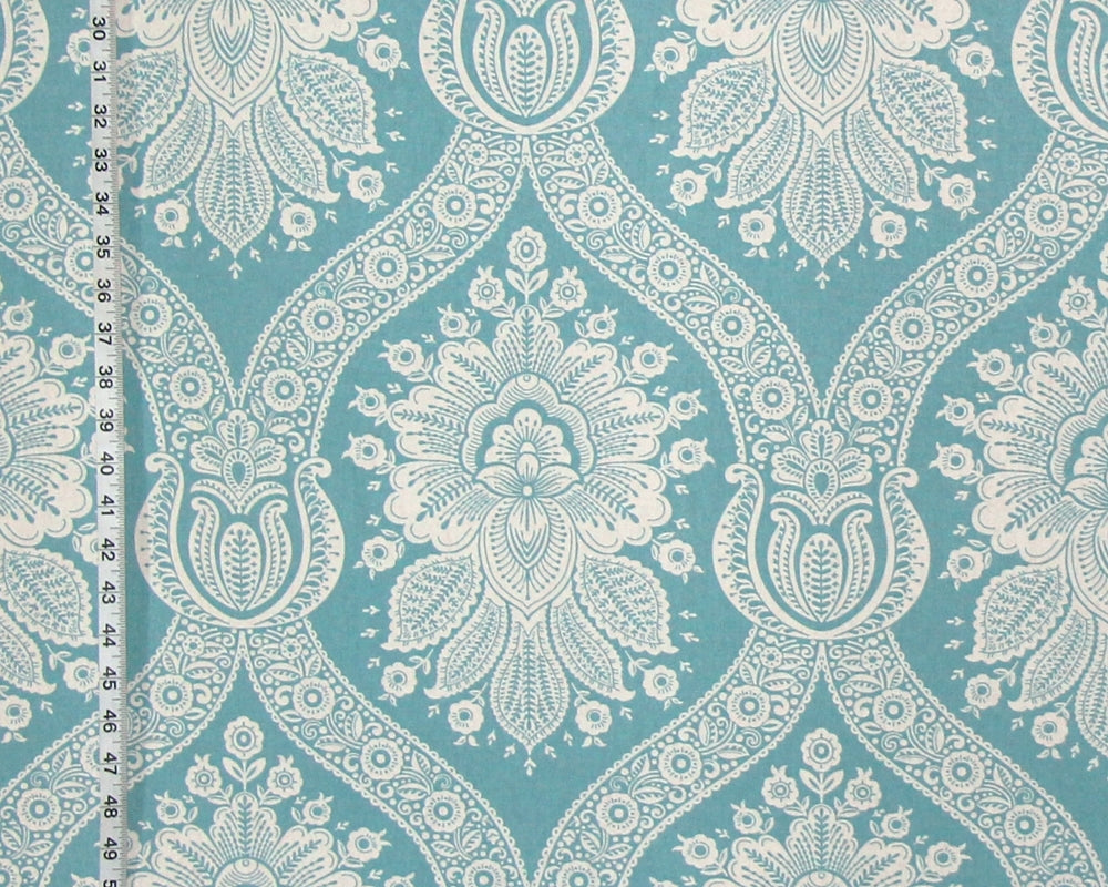 A close-up of a fabric or wallpaper pattern featuring a repeating white damask-style floral and ornamental motif on a turquoise blue background, with a ruler visible along the left edge for scale.