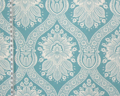 A close-up of a fabric or wallpaper pattern featuring a repeating white damask-style floral and ornamental motif on a turquoise blue background, with a ruler visible along the left edge for scale.