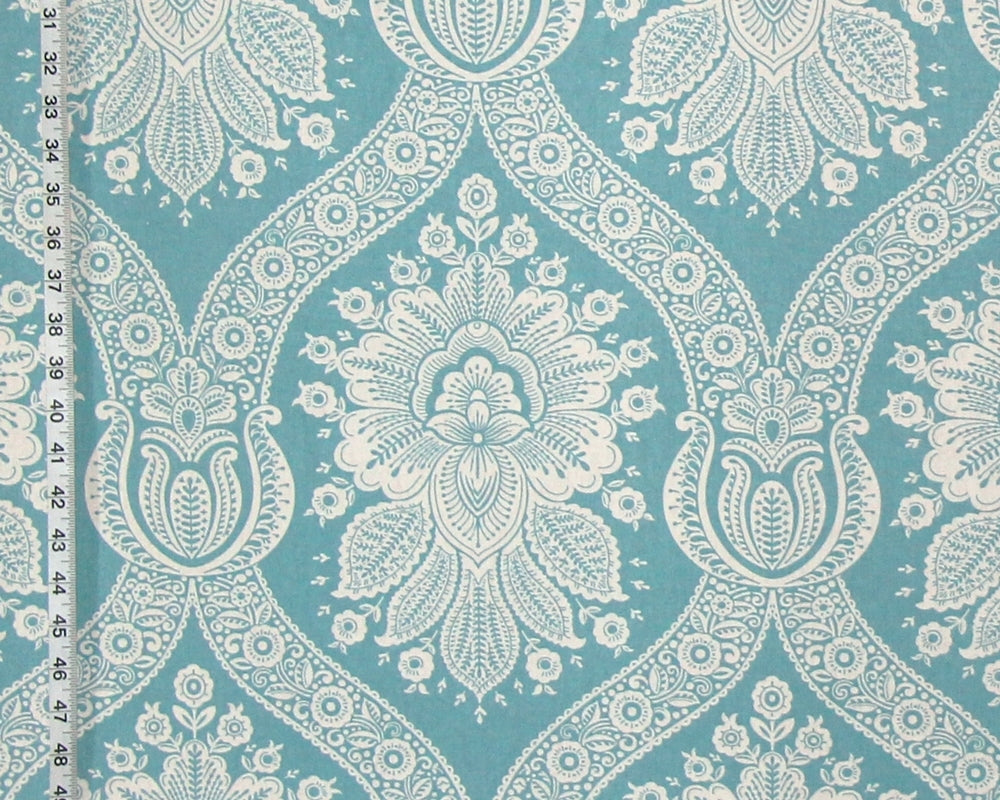 A close-up of a fabric or wallpaper pattern featuring a repeating white floral and paisley design on a turquoise blue background, with a white ruler visible along the left edge for scale.