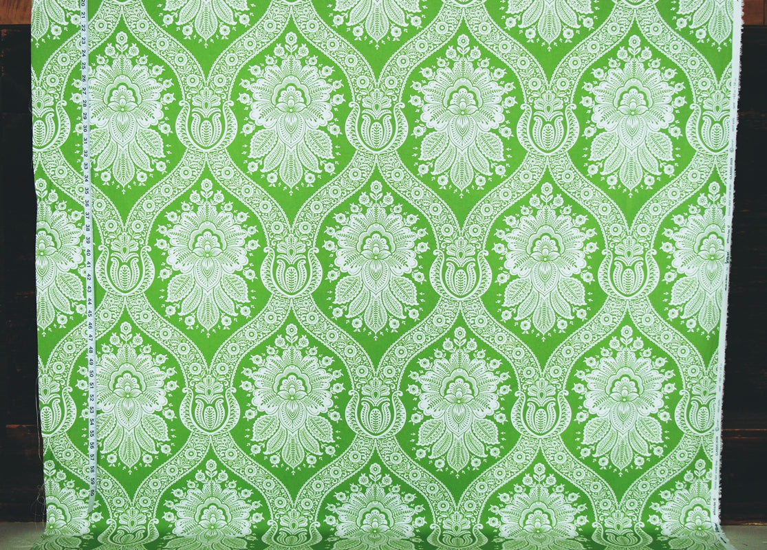Green and white damask patterned fabric hanging flat, showing a repeating ornate floral and scroll motif.