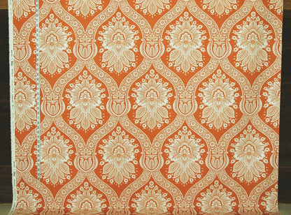 A close-up of patterned fabric with a repeating ornate damask-style floral motif in warm orange and off-white/cream.