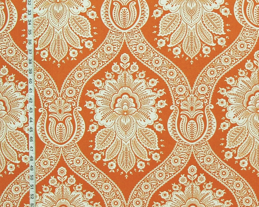 A close-up of a fabric or wallpaper pattern featuring a bold orange background with a cream-colored ornate damask floral design. A white measuring tape is visible along the left edge for scale.
