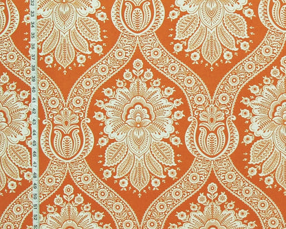 A close-up of a fabric or wallpaper pattern featuring a bold orange background with a cream-colored ornate damask floral design. A white measuring tape is visible along the left edge for scale.