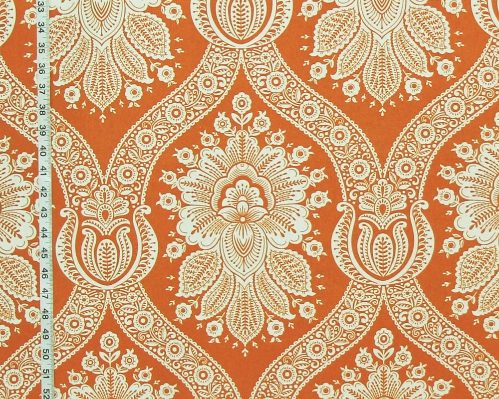 A close-up of a fabric pattern featuring a bold orange background with a detailed cream-colored paisley and floral damask design. A black and white fabric measuring tape is visible along the left edge to indicate scale.