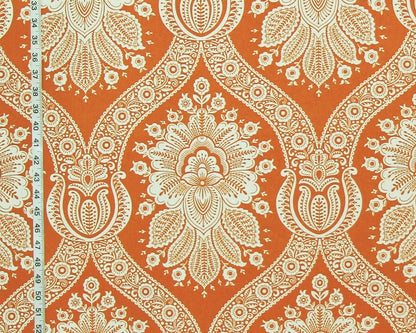 A close-up of a fabric pattern featuring a bold orange background with a detailed cream-colored paisley and floral damask design. A black and white fabric measuring tape is visible along the left edge to indicate scale.