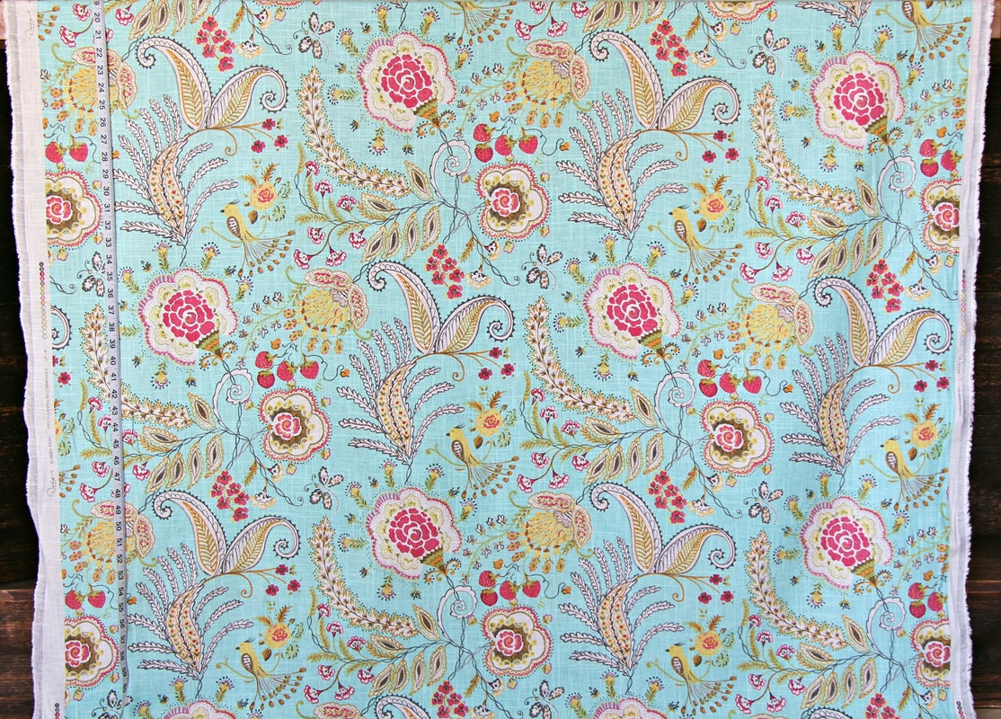 A close-up of a light blue fabric with an intricate hand-painted floral and paisley pattern in pink, yellow, green, and beige, finished with white fringed edges.