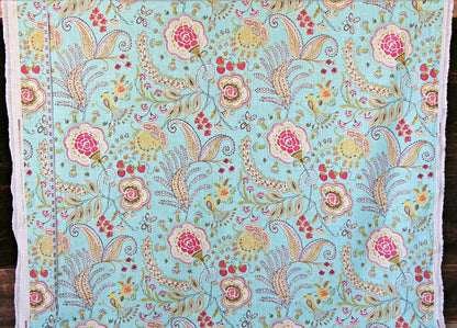 A close-up of a light blue fabric with an intricate hand-painted floral and paisley pattern in pink, yellow, green, and beige, finished with white fringed edges.