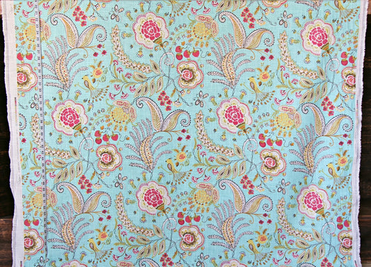 A close-up of a light blue fabric with an intricate hand-painted floral and paisley pattern in pink, yellow, green, and beige, finished with white fringed edges.