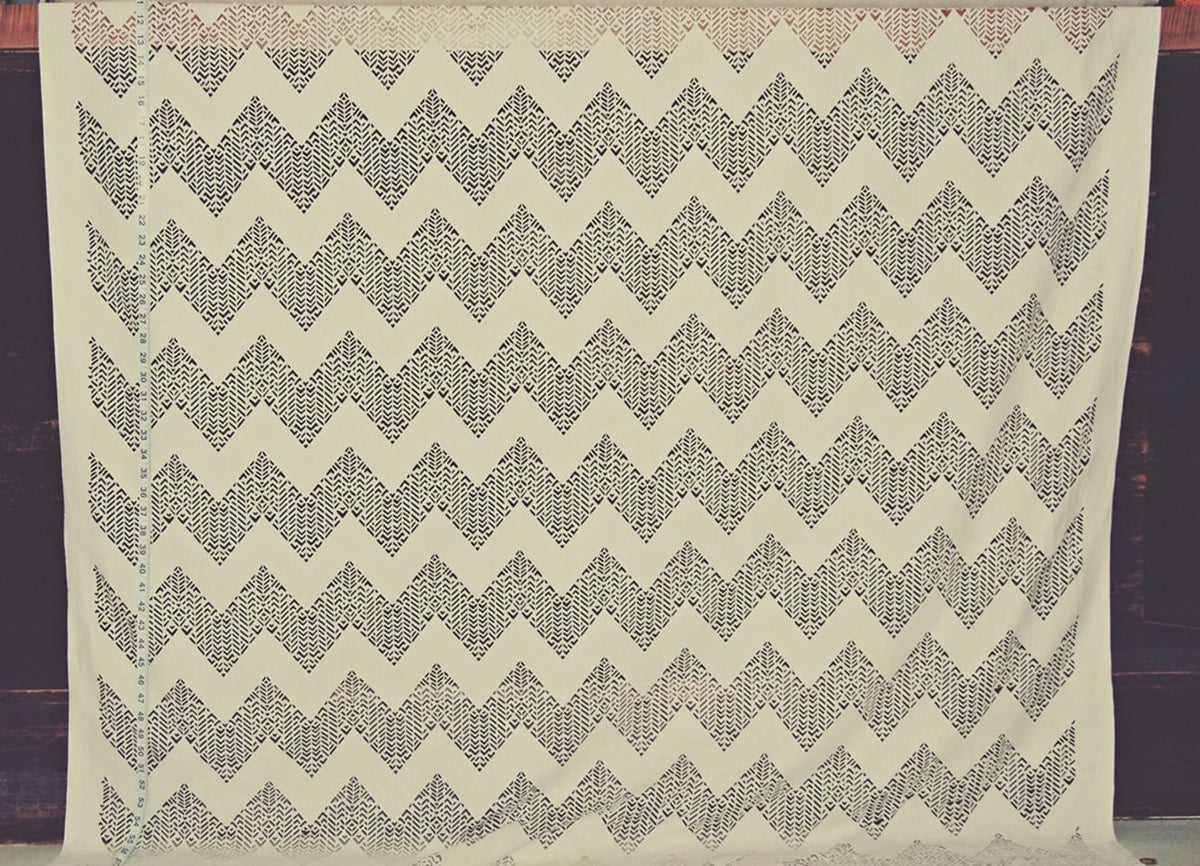 A square cream-colored fabric featuring a repeating dark gray geometric chevron pattern, displayed flat against a dark wooden background.
