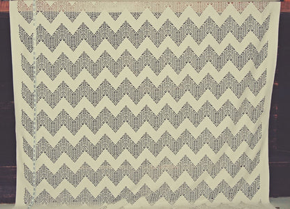 A square cream-colored fabric featuring a repeating dark gray geometric chevron pattern, displayed flat against a dark wooden background.
