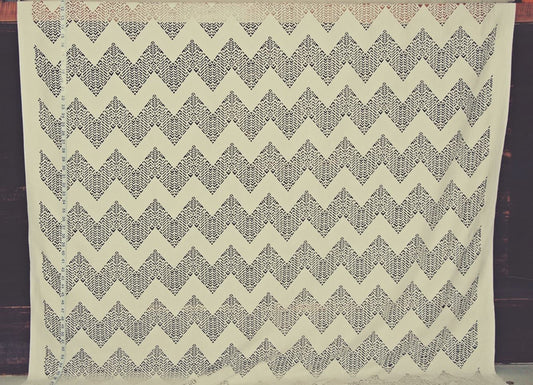 A square cream-colored fabric featuring a repeating dark gray geometric chevron pattern, displayed flat against a dark wooden background.