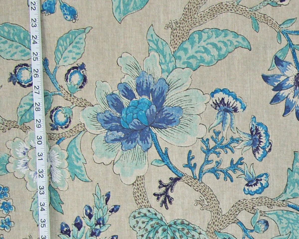 A close-up of a floral fabric print with large blue and white flowers, teal leaves, and brown branches on a light beige background, with a measuring tape visible along the left edge.