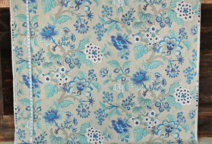 A close-up of a woven textile with an intricate floral pattern in shades of blue, teal, and white on a warm beige background, laid flat on a dark wood surface.