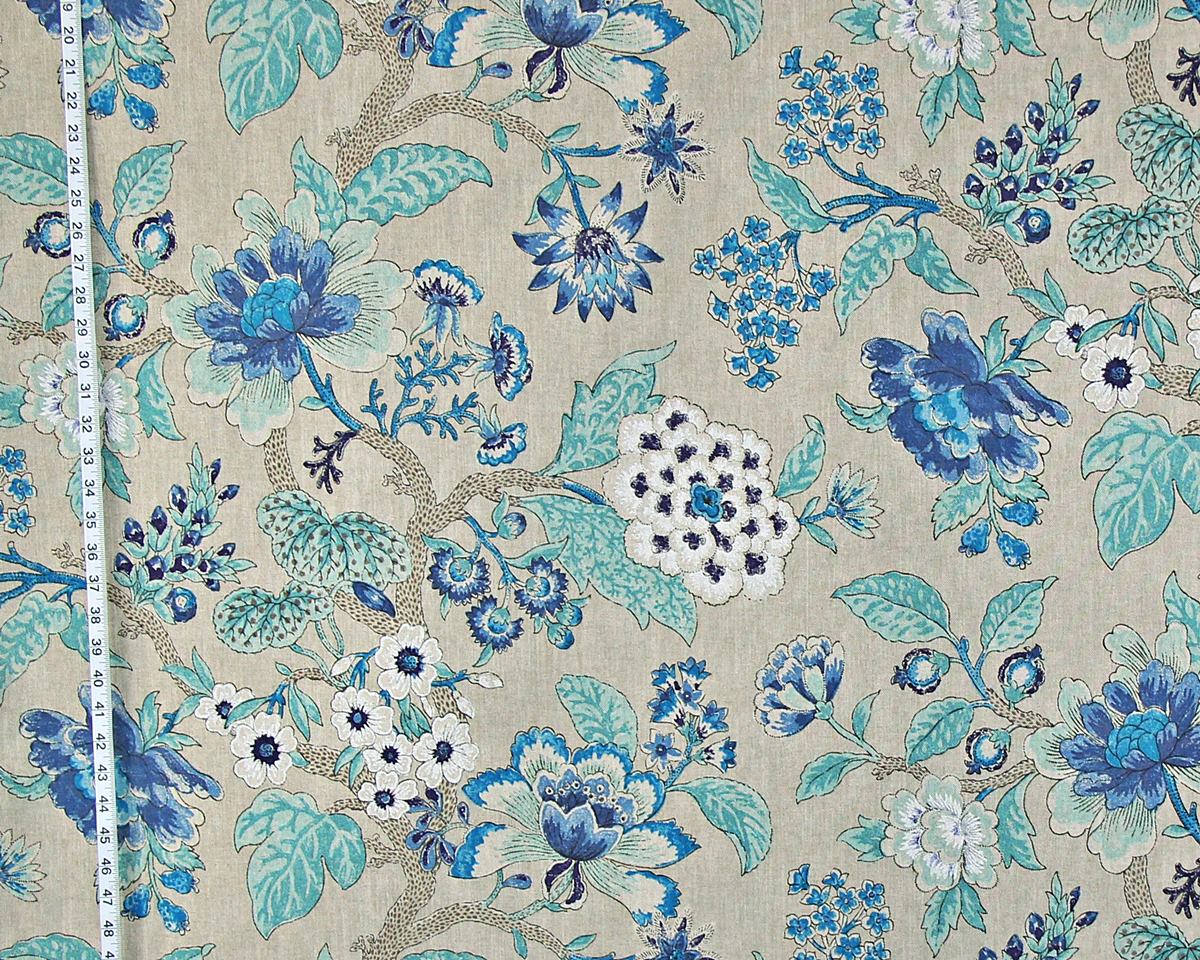 A close-up of a floral fabric with a repeating pattern of blue and teal flowers, white blossoms, and beige branches on a light beige background, with a white measuring tape visible along the left edge.