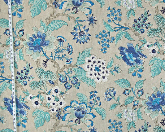 A close-up of a floral fabric with a repeating pattern of blue and teal flowers, white blossoms, and beige branches on a light beige background, with a white measuring tape visible along the left edge.