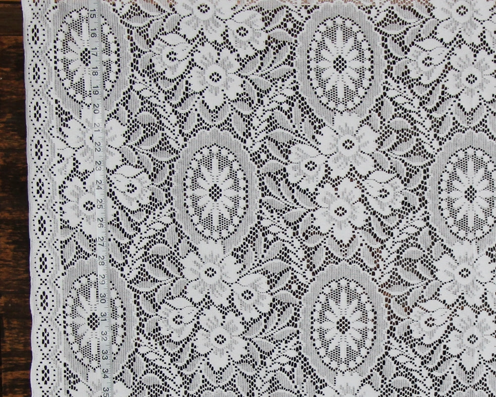 White floral lace fabric with intricate embroidered patterns and a scalloped border, laid flat on a dark wooden surface.