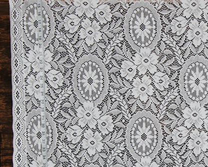 White floral lace fabric with intricate embroidered patterns and a scalloped border, laid flat on a dark wooden surface.